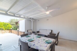 Un patio cubierto con una mesa para cuatro, rodeado de sillas de mimbre, un ventilador de techo y una pared de piedra al fondo.
