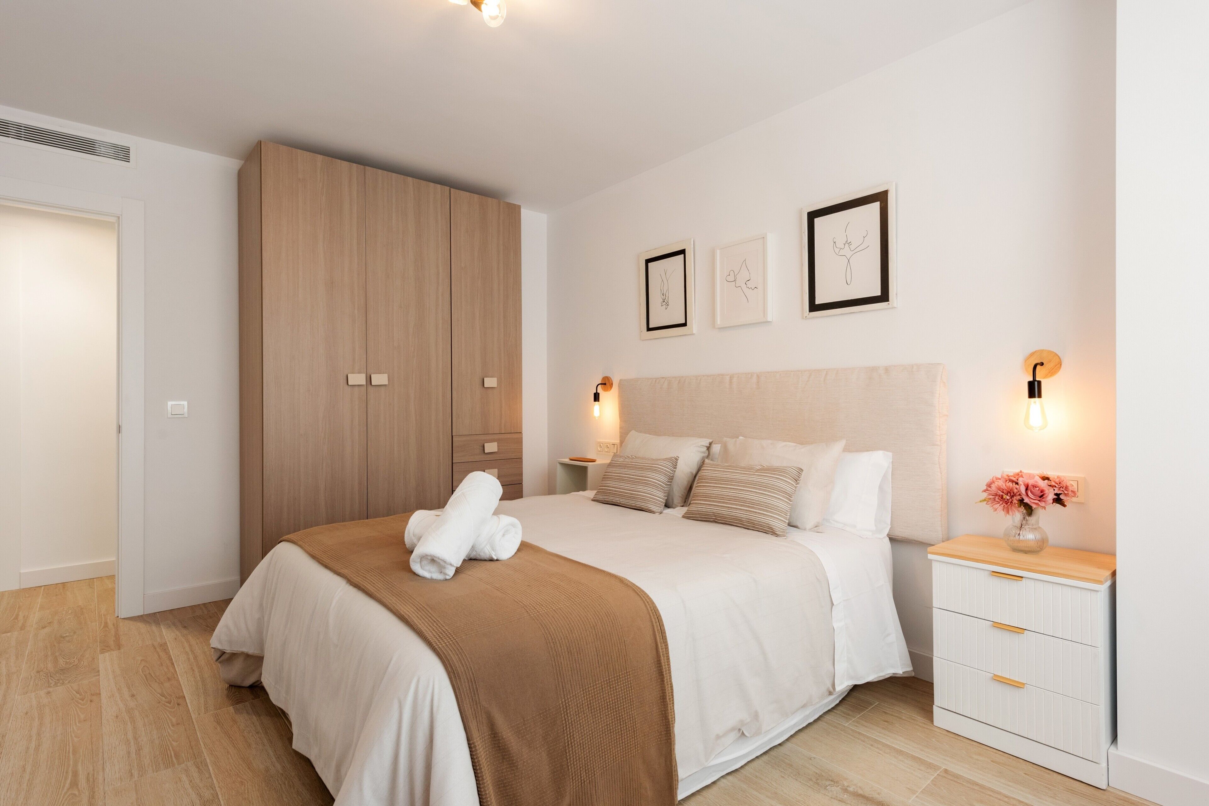 A bedroom with a bed, a wooden wardrobe, a bedside table with a lamp, and wall art.