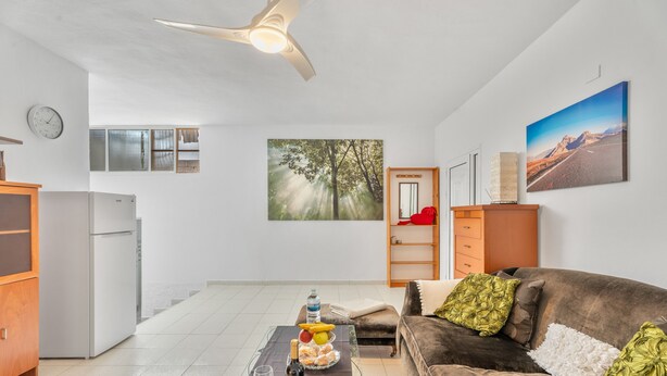 Una sala de estar con un sofá marrón, una mesa de centro de vidrio, una mesa de comedor con fruteros, una botella de vino y un reloj en la pared.
