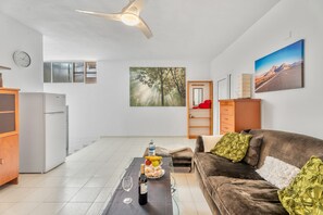 A living room with a brown sofa, a glass coffee table, a dining table with a fruit bowl, a bottle of wine, and a clock on the wall.