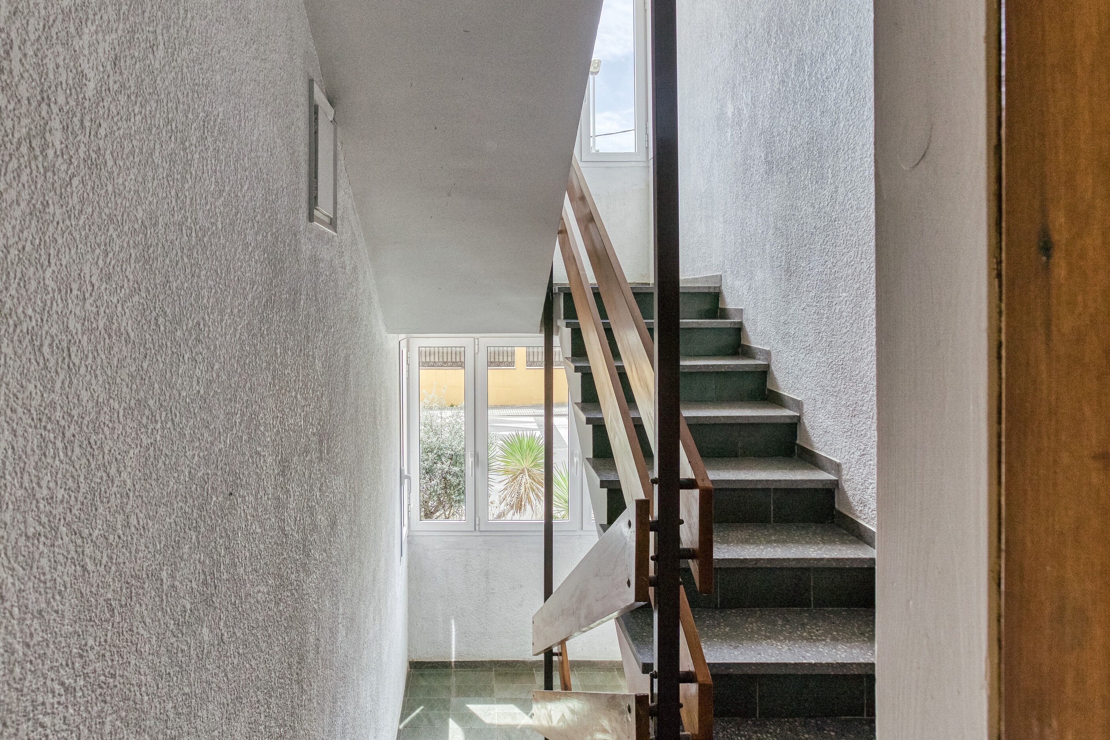 A staircase with wooden handrails leading to an upper floor, a window with a view of greenery, and a textured wall.
