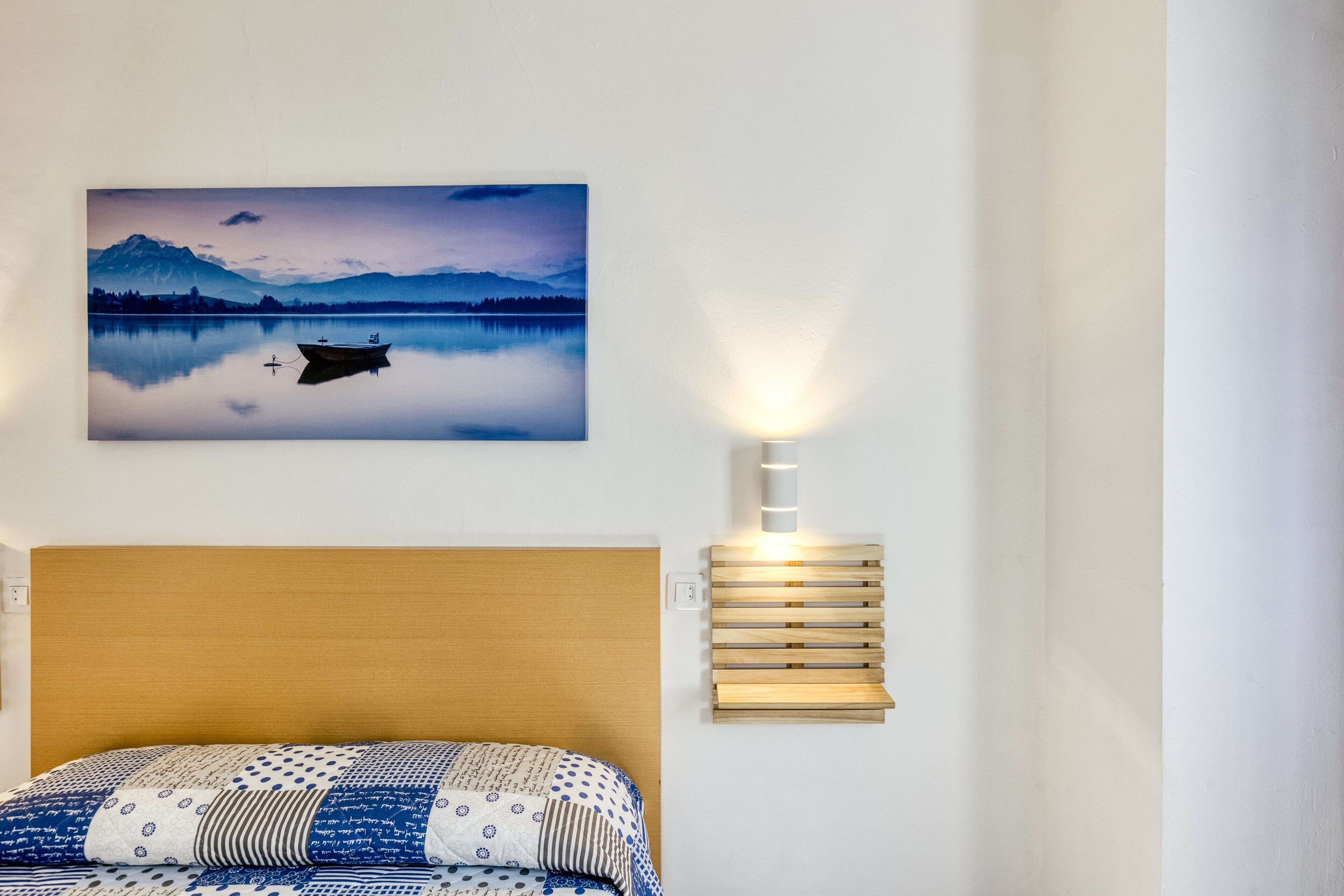 A bedroom with a bed, a wall-mounted light, and a framed landscape painting.