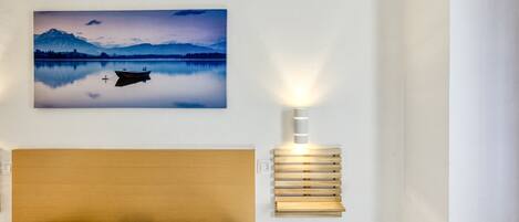 A bedroom with a bed, a wall-mounted light, and a framed landscape painting.