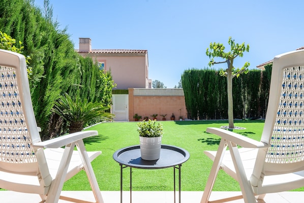 Un patio con dos sillas blancas, una pequeña mesa redonda con una planta en maceta y un césped bien cuidado.