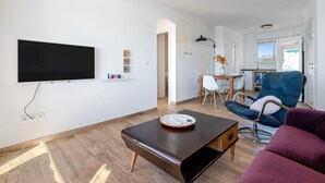 Un salon moderne doté d’un téléviseur à écran plat, d’une table basse, d’un canapé, d’un coin repas et d’une cuisine visible à travers une porte ouverte.