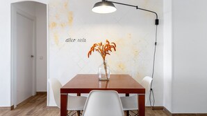 Un coin repas avec une table en bois, des chaises blanches, un vase de fleurs et un mur orné d’un motif texturé et d’un texte.
