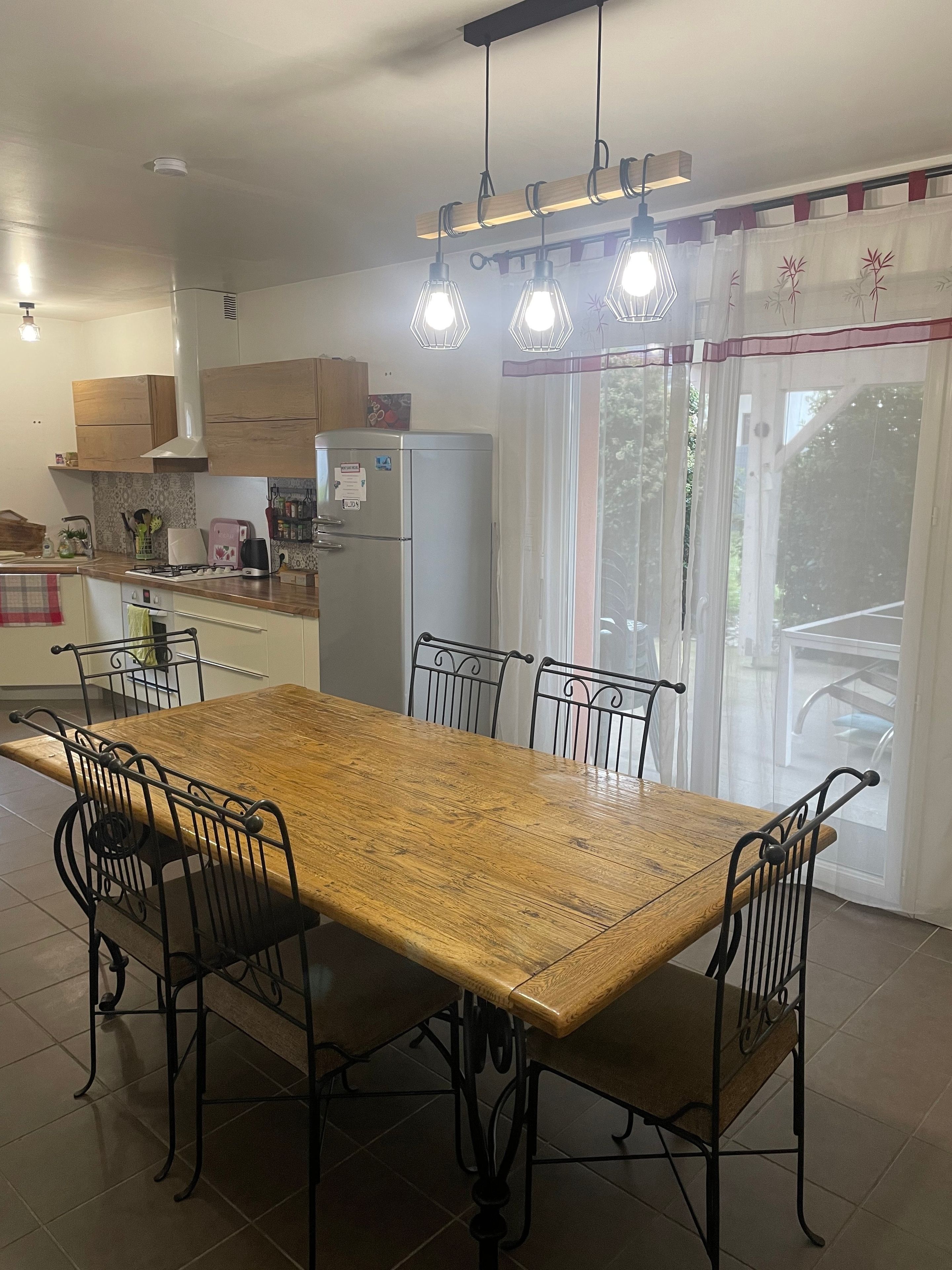 A kitchen with a wooden table and chairs, a refrigerator, and a window with curtains.