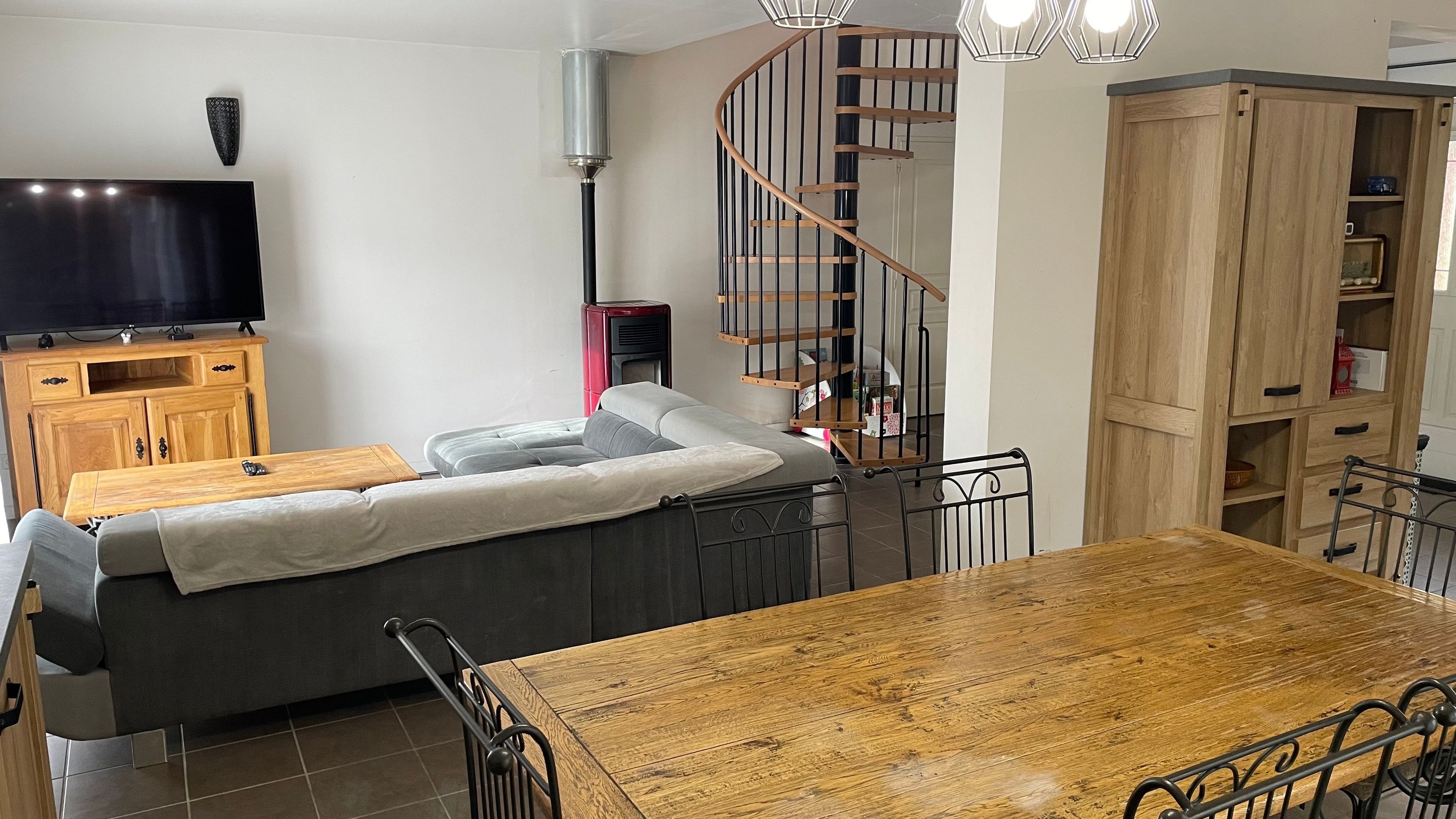 A living room with a sofa, a wooden dining table, a TV on a wooden cabinet, a spiral staircase, and a wooden wardrobe.