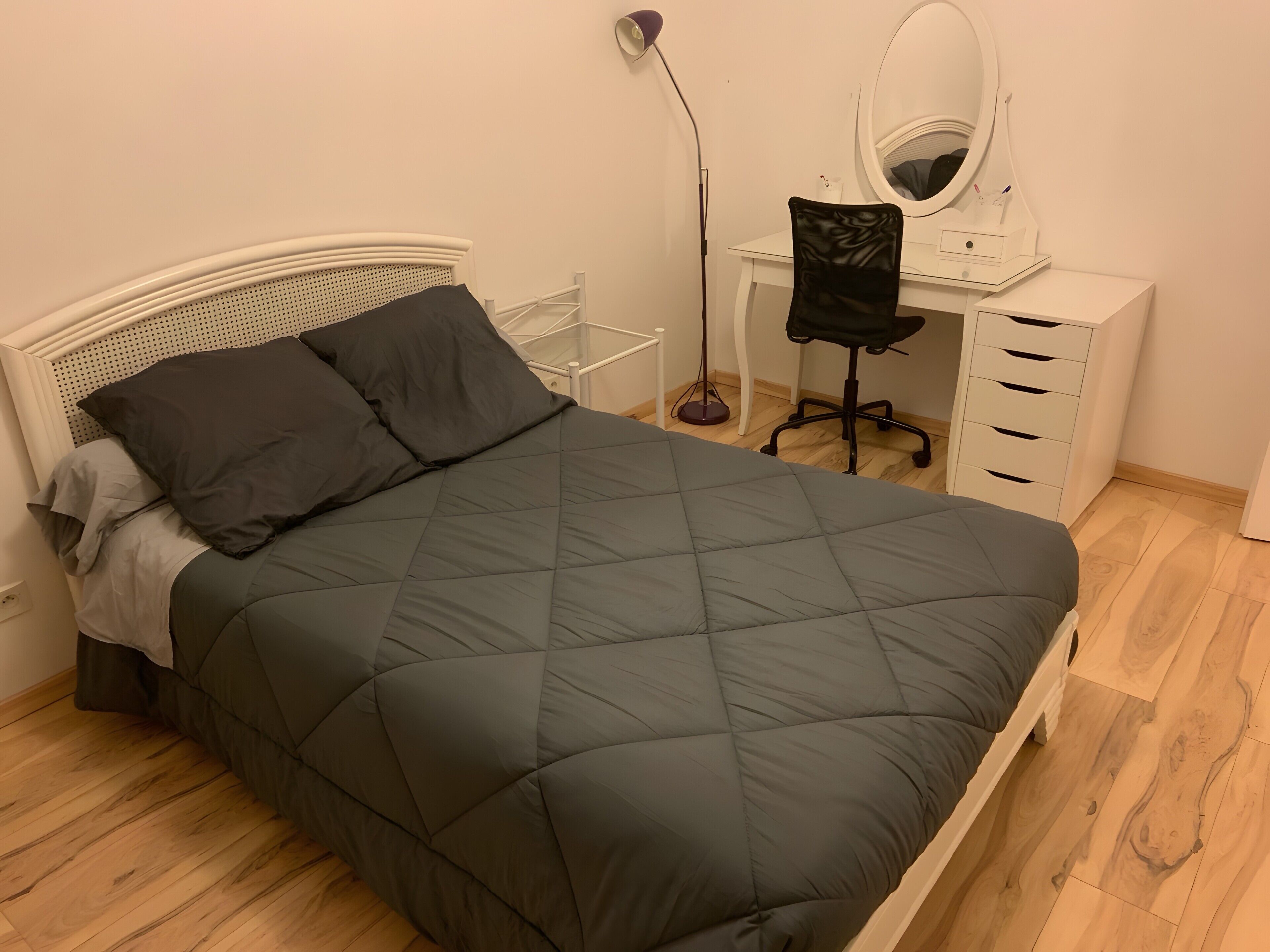 Un lit bien fait, recouvert d’une couverture sombre, une table de chevet blanche, une chaise de bureau noire, un bureau avec un miroir et une lampe sur pied.