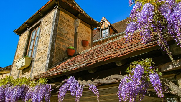 Un edificio de piedra con techo inclinado, una ventana y flores moradas colgantes.