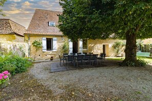 Un bâtiment en pierre avec un toit en tuiles, une table entourée de chaises et un arbre au premier plan.