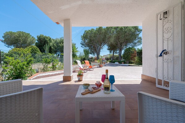 Un patio exterior con sillas de mimbre, una mesa con comida y bebidas, y vistas a árboles y una carretera.
