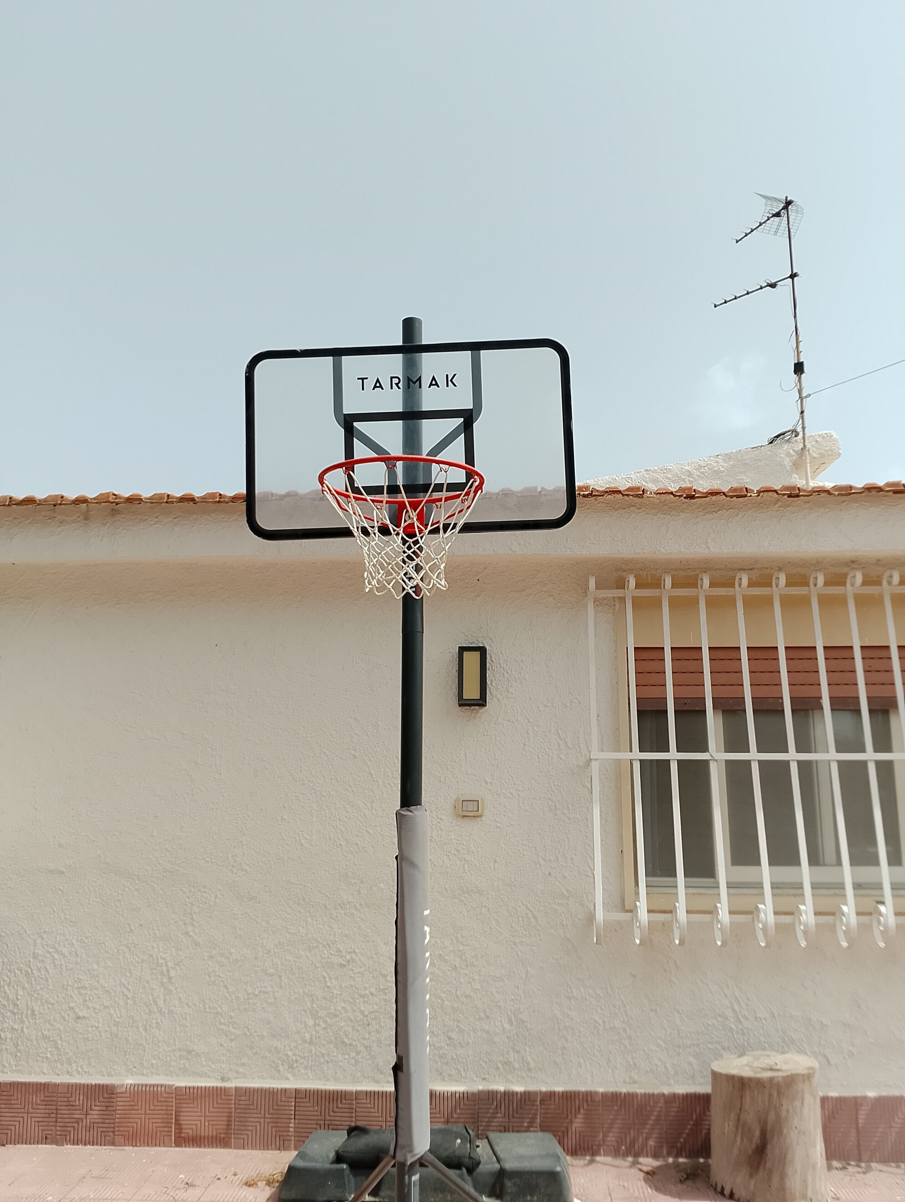 Un panier de basket avec une enseigne TARMAK, une fenêtre avec des barreaux et un poteau en bois.