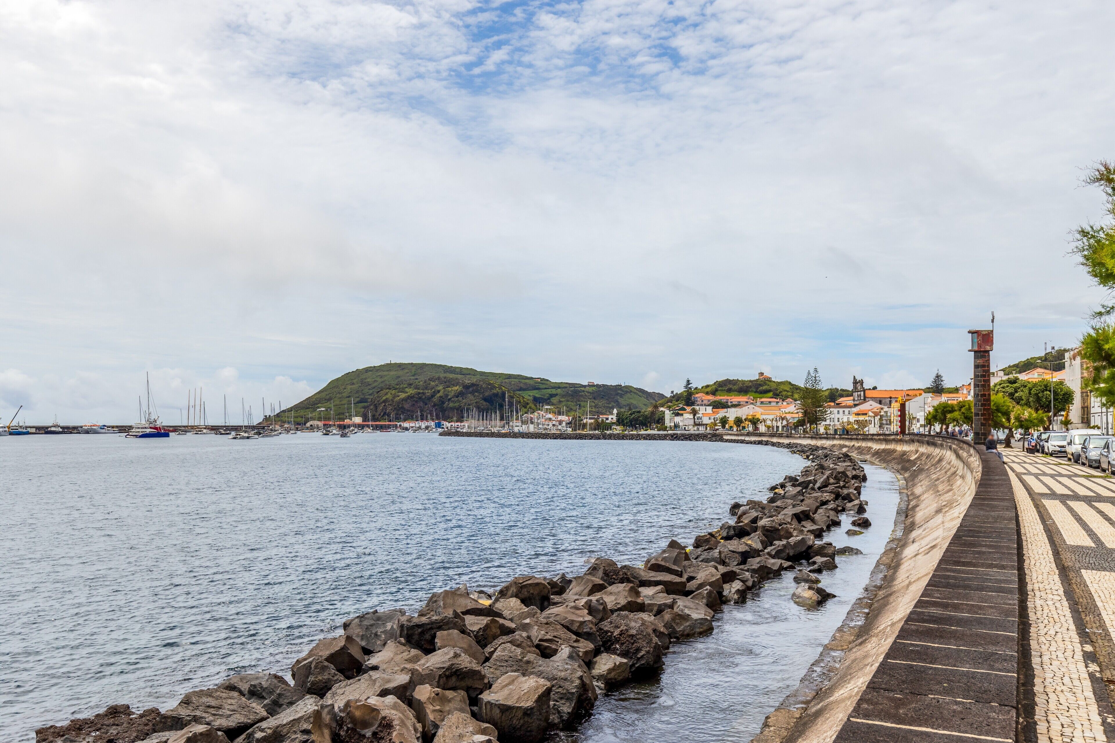 A coastal promenade with a stone barrier, a clear sky, and distant buildings.