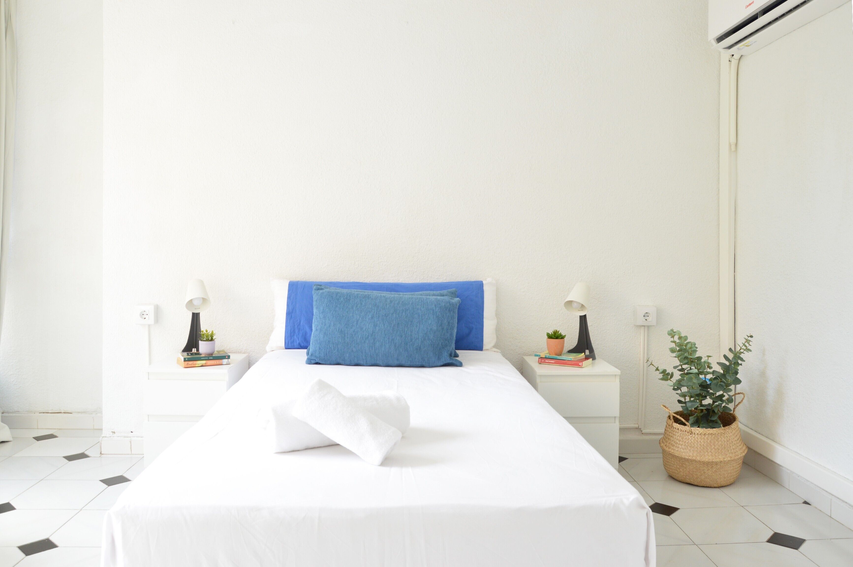 A neatly made bed with a blue pillow, white bedding, two bedside tables with lamps and books, and a potted plant.