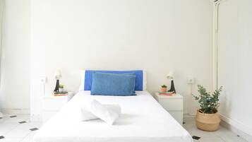 A neatly made bed with a blue pillow, white bedding, two bedside tables with lamps and books, and a potted plant.