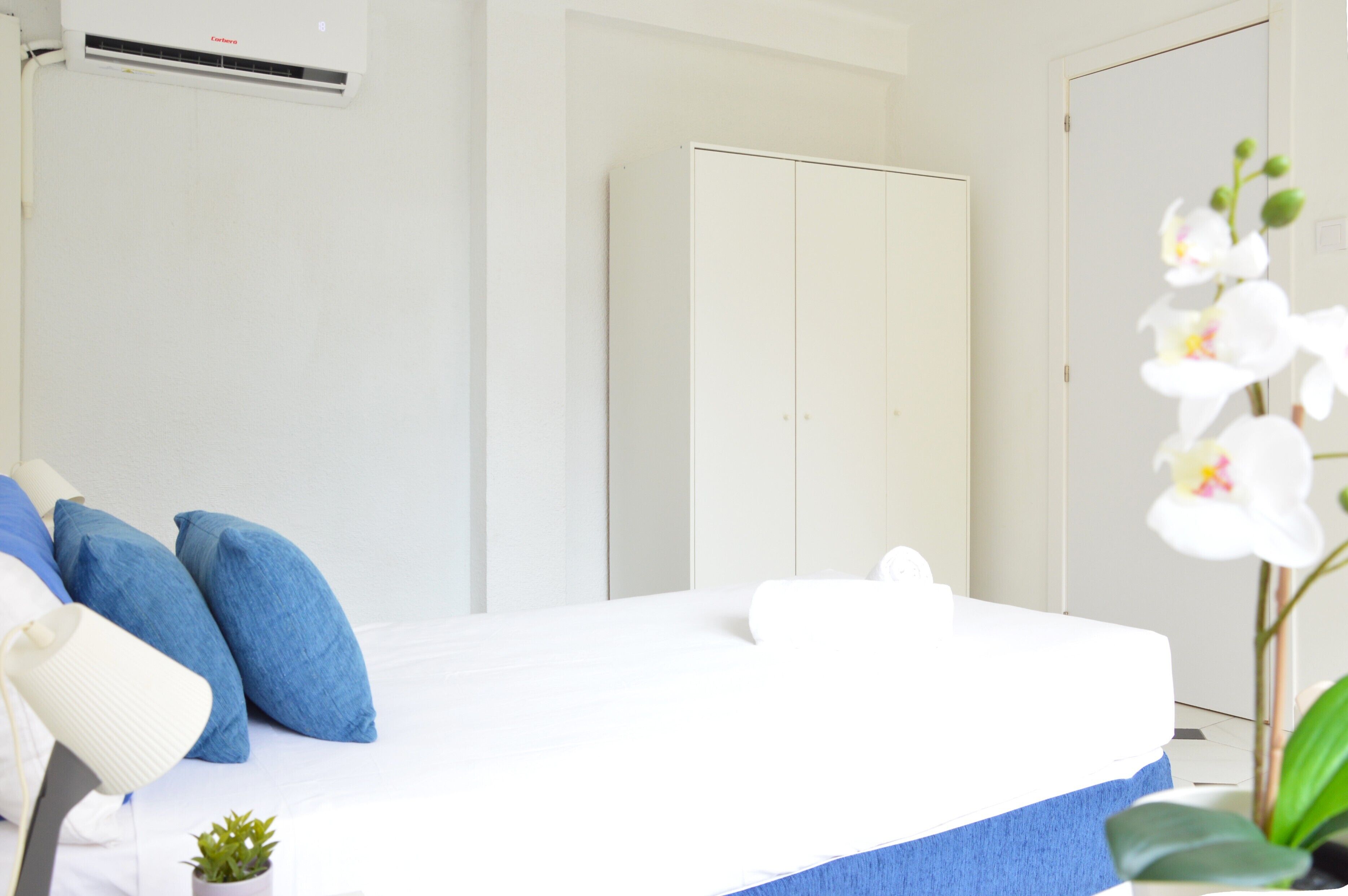 A bedroom with a bed, pillows, a small plant, an air conditioner, and a wardrobe.