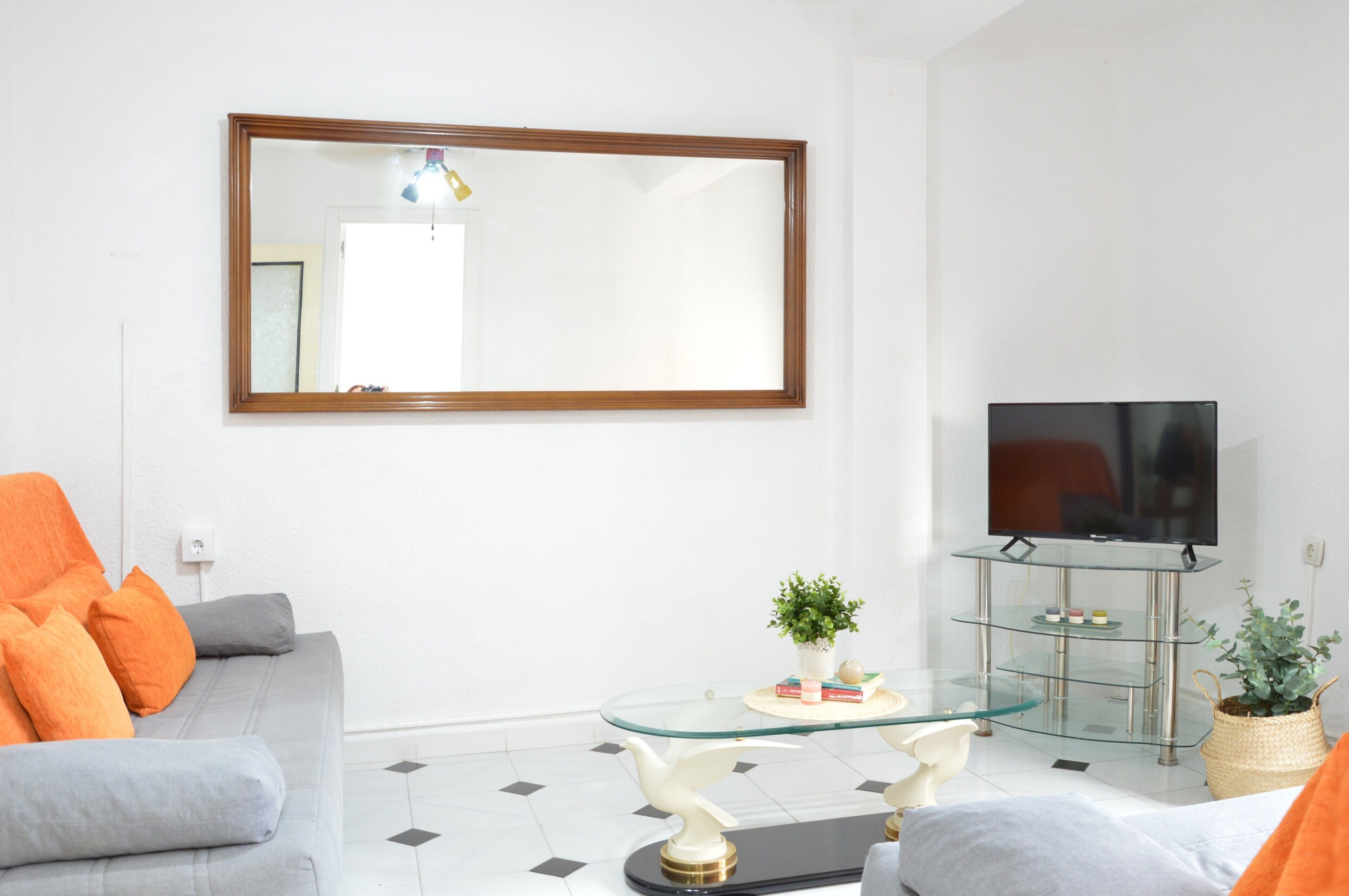 Un salon avec un canapé, une table basse en verre, un téléviseur posé sur un meuble et un miroir accroché au mur.
