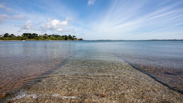 Un lac paisible aux eaux limpides, une rive lointaine bordée d’arbres et de bâtiments, et un ciel dégagé.