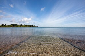 Un lac paisible aux eaux limpides, une rive lointaine bordée d’arbres et de bâtiments, et un ciel dégagé.