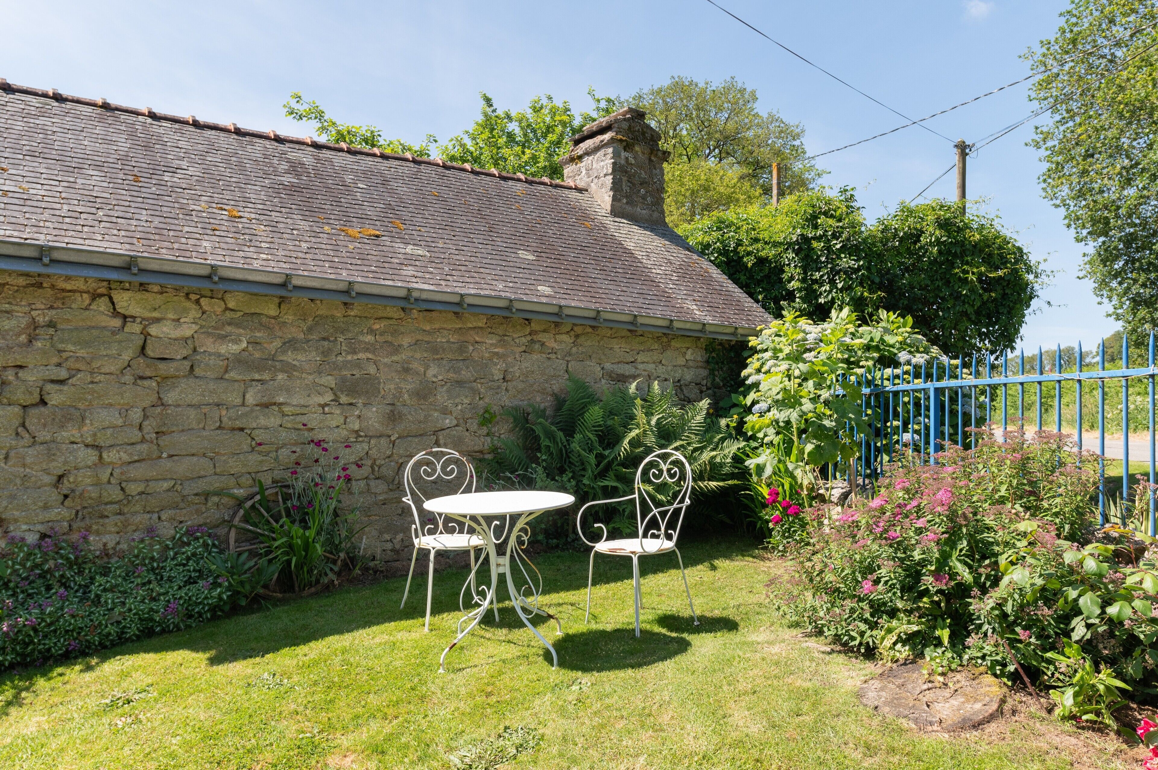 A stone building with a tiled roof, a small garden with a table and chairs, and a blue fence.