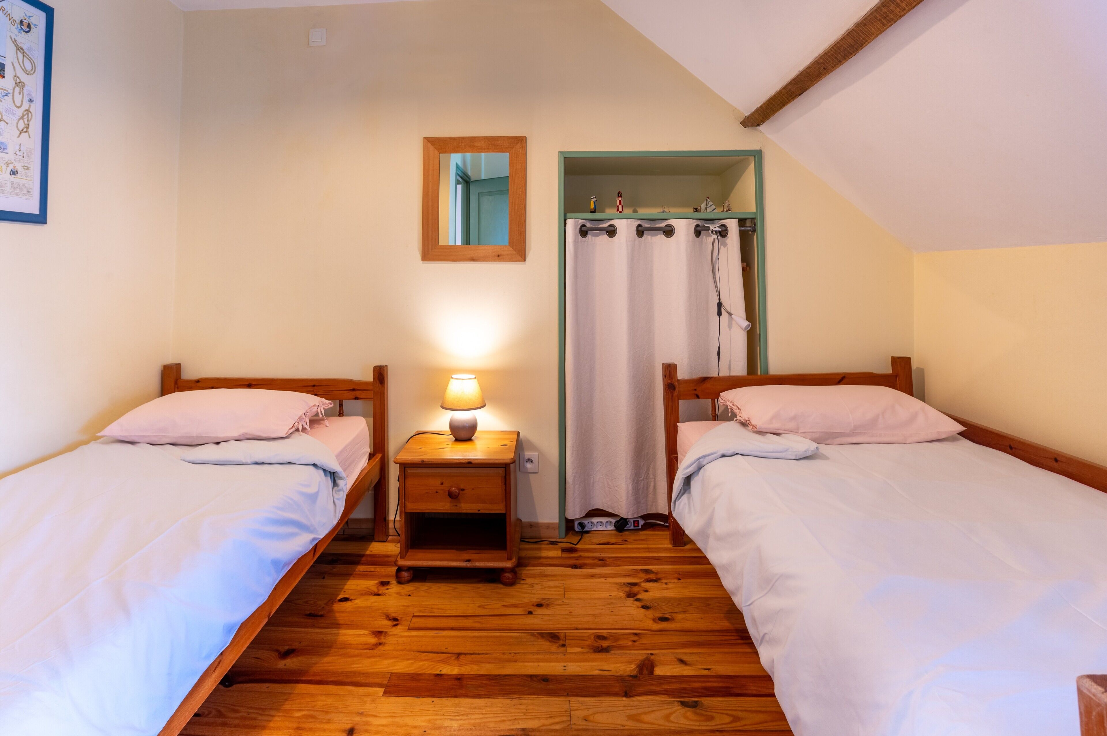 Quarto com duas camas de solteiro, mesa de cabeceira com abajur, piso de madeira, espelho e cortina de chuveiro.