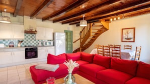 Un salon chaleureux avec un canapé rouge, des poutres en bois au plafond, une cuisine avec des meubles blancs et un escalier.