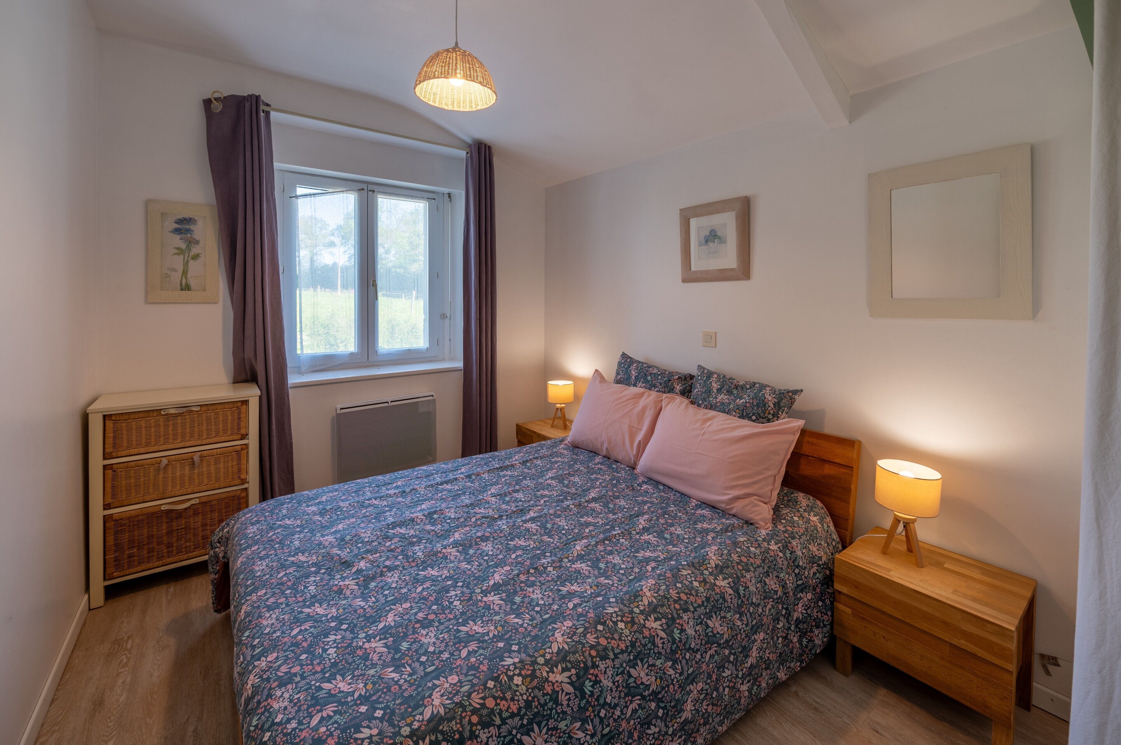 Une chambre à coucher comprenant un lit, des lampes de chevet, une commode, une fenêtre avec des rideaux et des œuvres d’art murales.