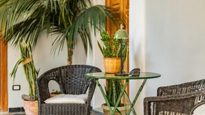 Una silla de mimbre, una mesita con una lámpara y plantas en macetas en una habitación con una puerta de madera y una pared blanca.