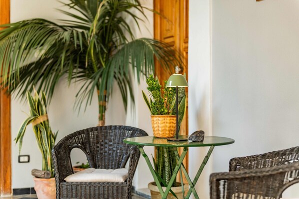 A wicker chair, a small table with a lamp, and potted plants in a room with a wooden door and a white wall.