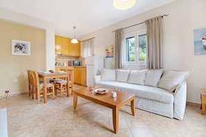 A living room with a sofa, dining table, and chairs, a kitchen area with cabinets and appliances, and a window with curtains.