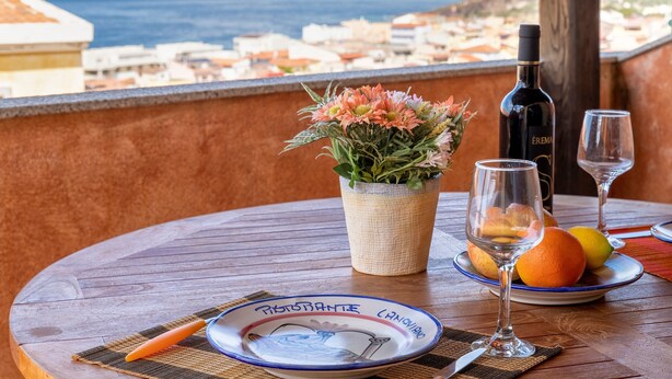 A table with a bottle of wine, glasses, a plate with fruit, and a vase of flowers on a balcony overlooking a coastal town. - Apartment 'Bella Vista Castelsardo' with Sea View, Balcony and Air Conditioning (Castelsardo)