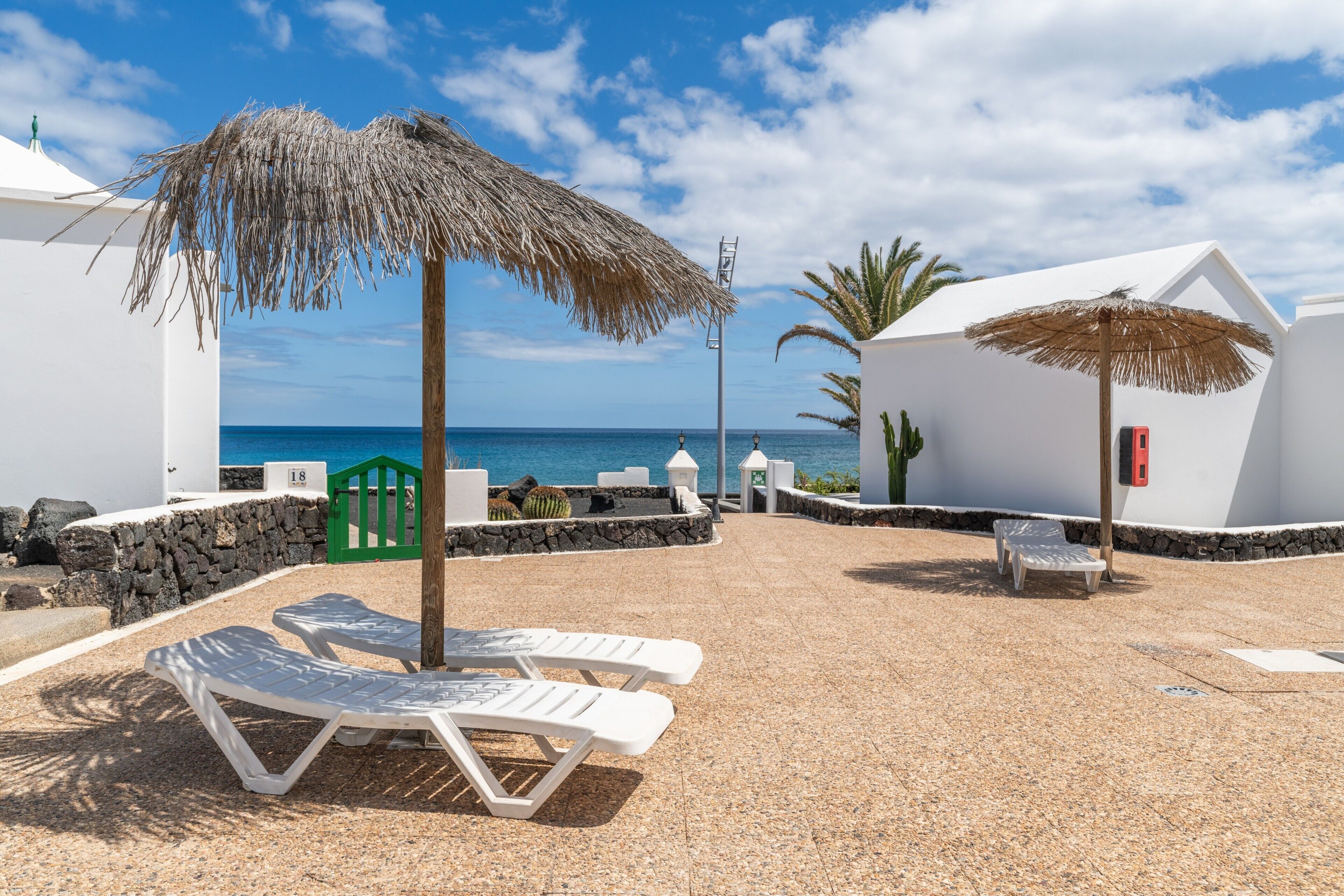 Een hotel aan het strand met witte muren, een rieten parasol en ligstoelen.