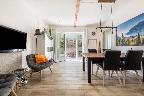 A modern living room with a flat-screen TV, a dining area with a wooden table and chairs, and a large window with a view of a rocky landscape.