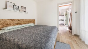 Ett sovrum med en sänggavel i trä, en säng med mönstrat sänglinne, en hylla med inramade bilder och en hall som leder till ett badrum.