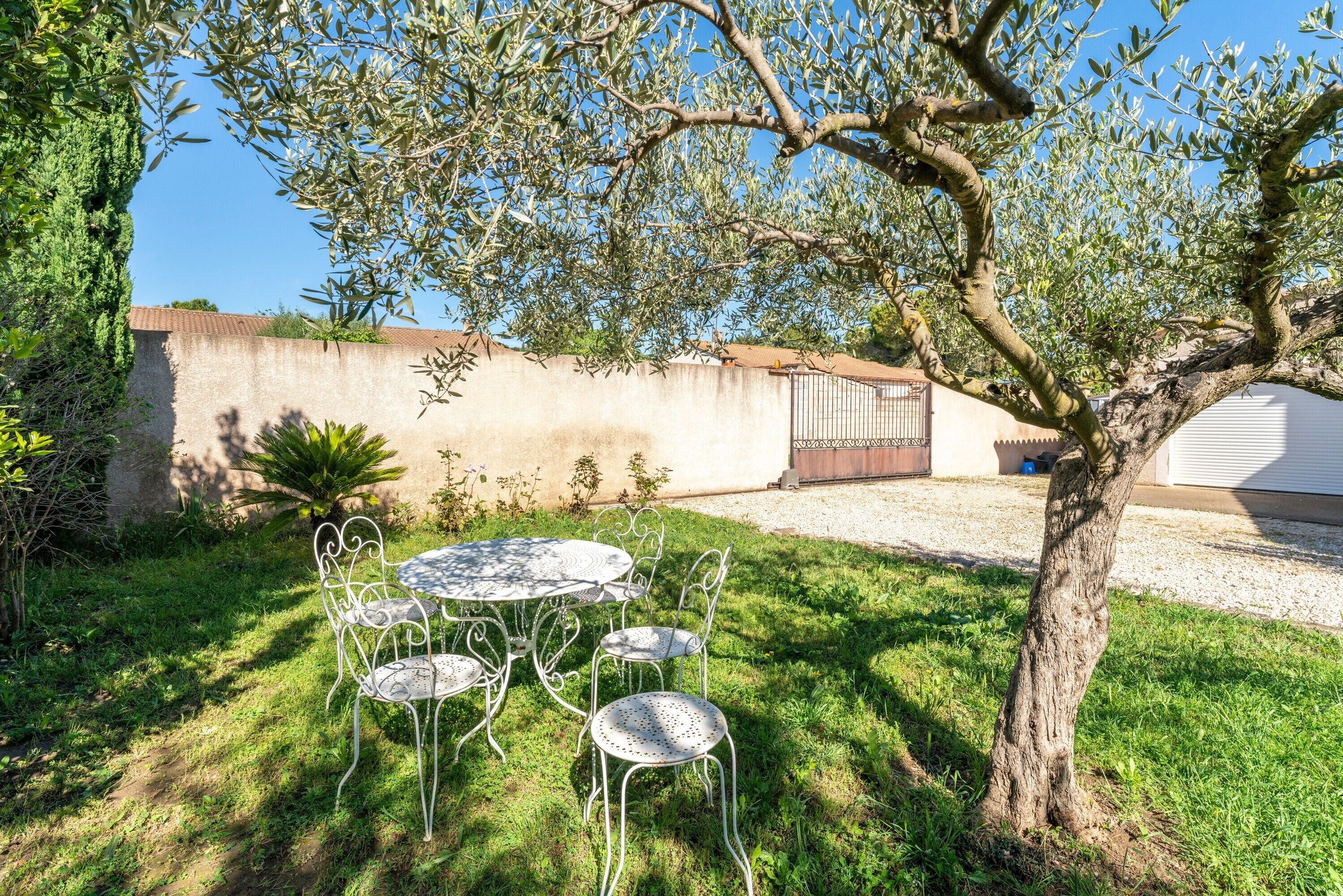 Un patio con una mesa y sillas de metal, rodeado de vegetación y un árbol.