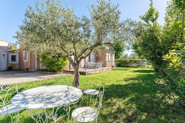 A courtyard with a white metal table and chairs, surrounded by olive trees and a house with a balcony.