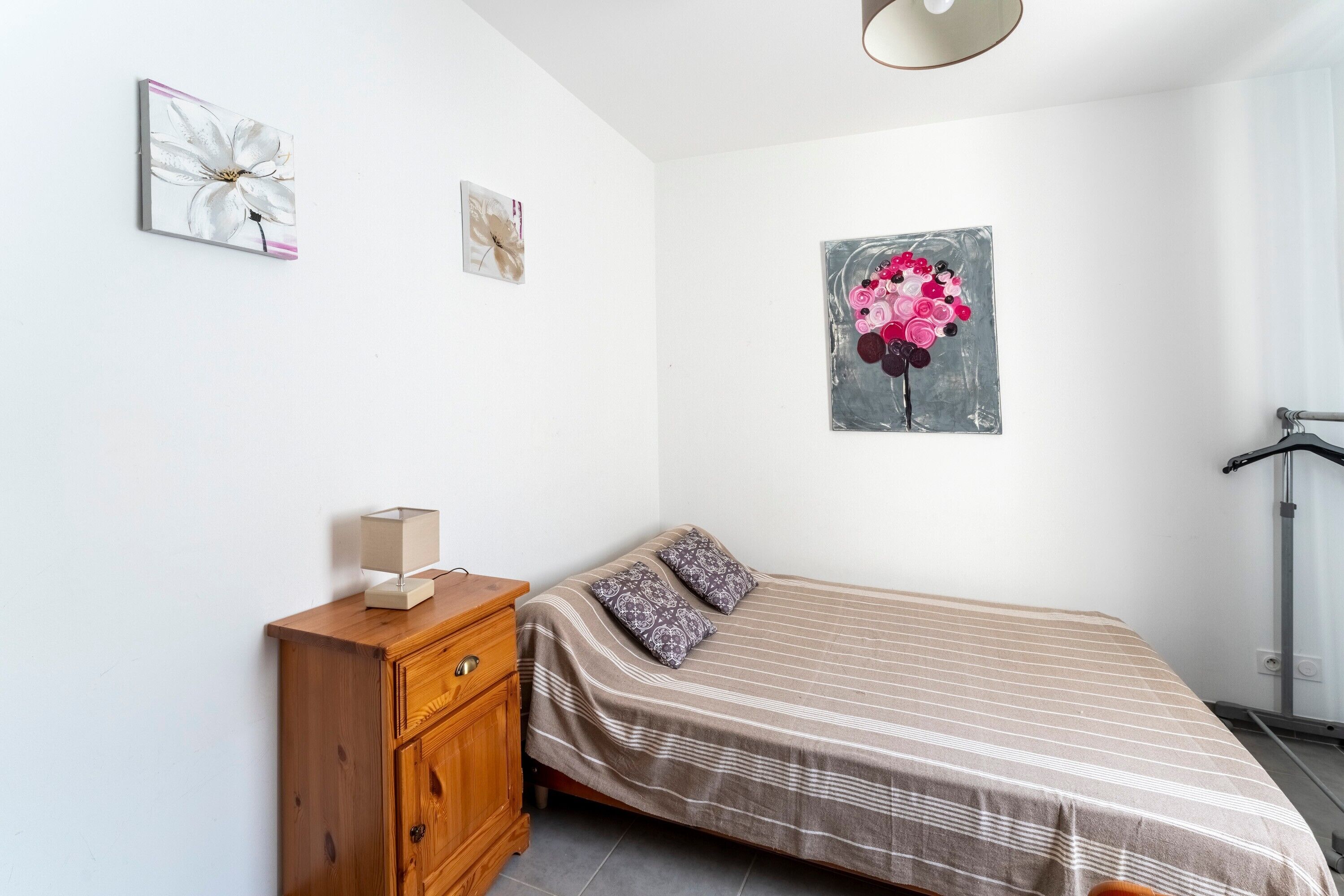 A bedroom with a bed, a wooden dresser, a wall-mounted lamp, and two paintings.