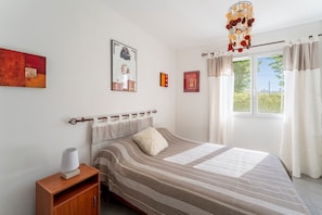 A bedroom with a bed, a bedside table, a lamp, a window with curtains, and wall art.