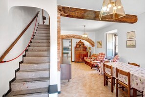 Un escalier avec une rampe rouge, une poutre en bois au plafond, un coin repas avec une table et des chaises, et une cuisine en arrière-plan.