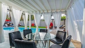 Een terras met een glazen tafel en stoelen, een zwembad en witte gordijnen.