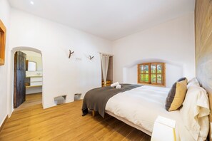 Une chambre avec un plancher en bois, un lit recouvert d’une couvre-lit blanche et grise, une fenêtre donnant sur un beau paysage et une porte en bois.