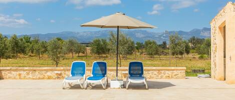 A pool area with three lounge chairs, a large umbrella, and a view of mountains.