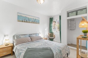 Un dormitorio con cama, mesitas de noche, lámpara, silla, un cuadro y una ventana con cortinas.