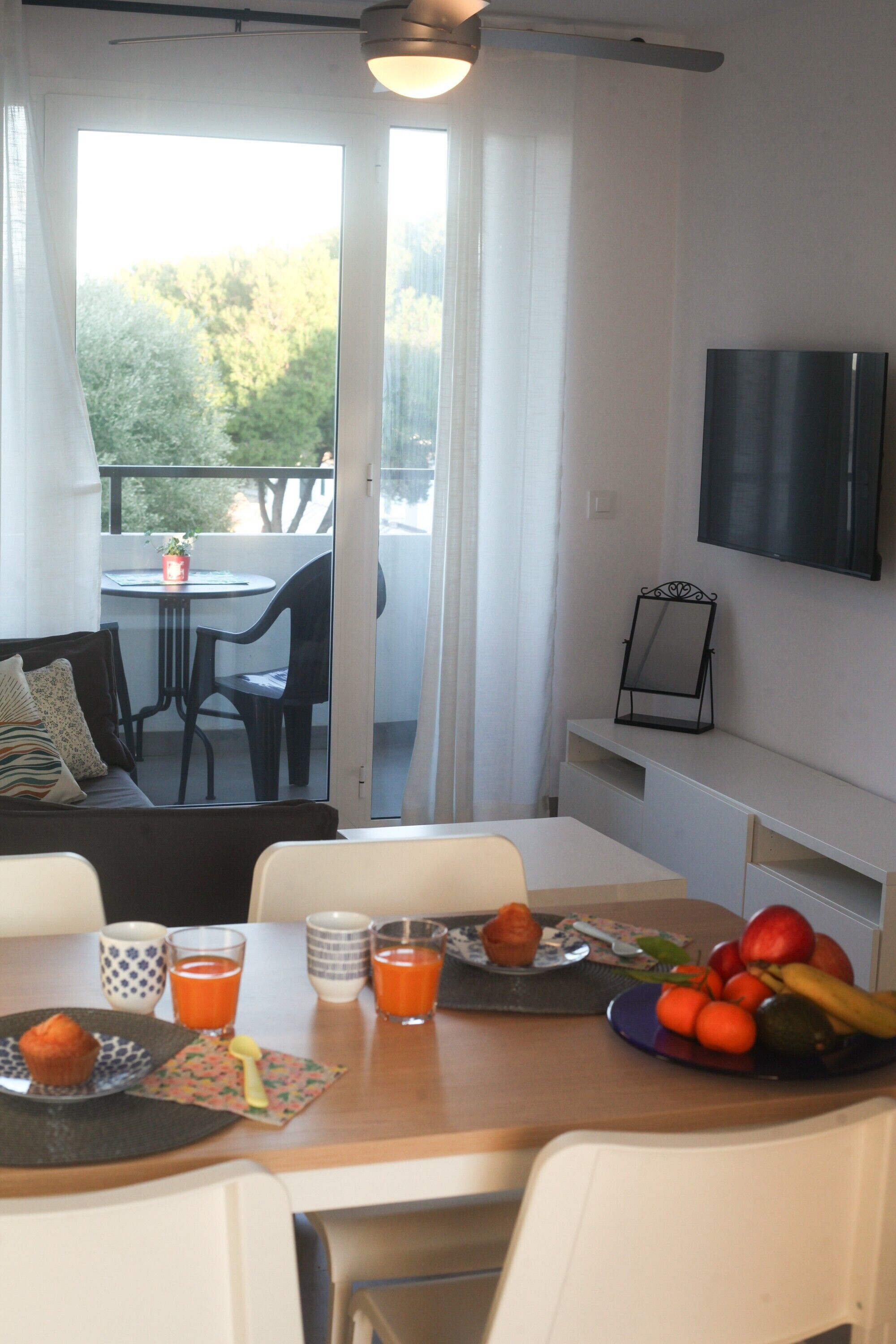 Un coin repas avec une table dressée pour deux personnes, un canapé, un téléviseur fixé au mur et un balcon avec une table et des chaises.