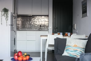 Una cocina moderna con armarios blancos, un comedor con sillas blancas y un sofá de color oscuro con un cojín estampado.