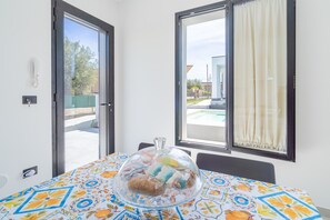 A room with a table covered by a colorful tablecloth, a glass dome over food, a sliding glass door, and a window with a curtain.