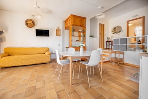 A modern living room with a yellow sofa, a round dining table with chairs, a kitchen area with wooden cabinets, and a TV mounted on the wall.