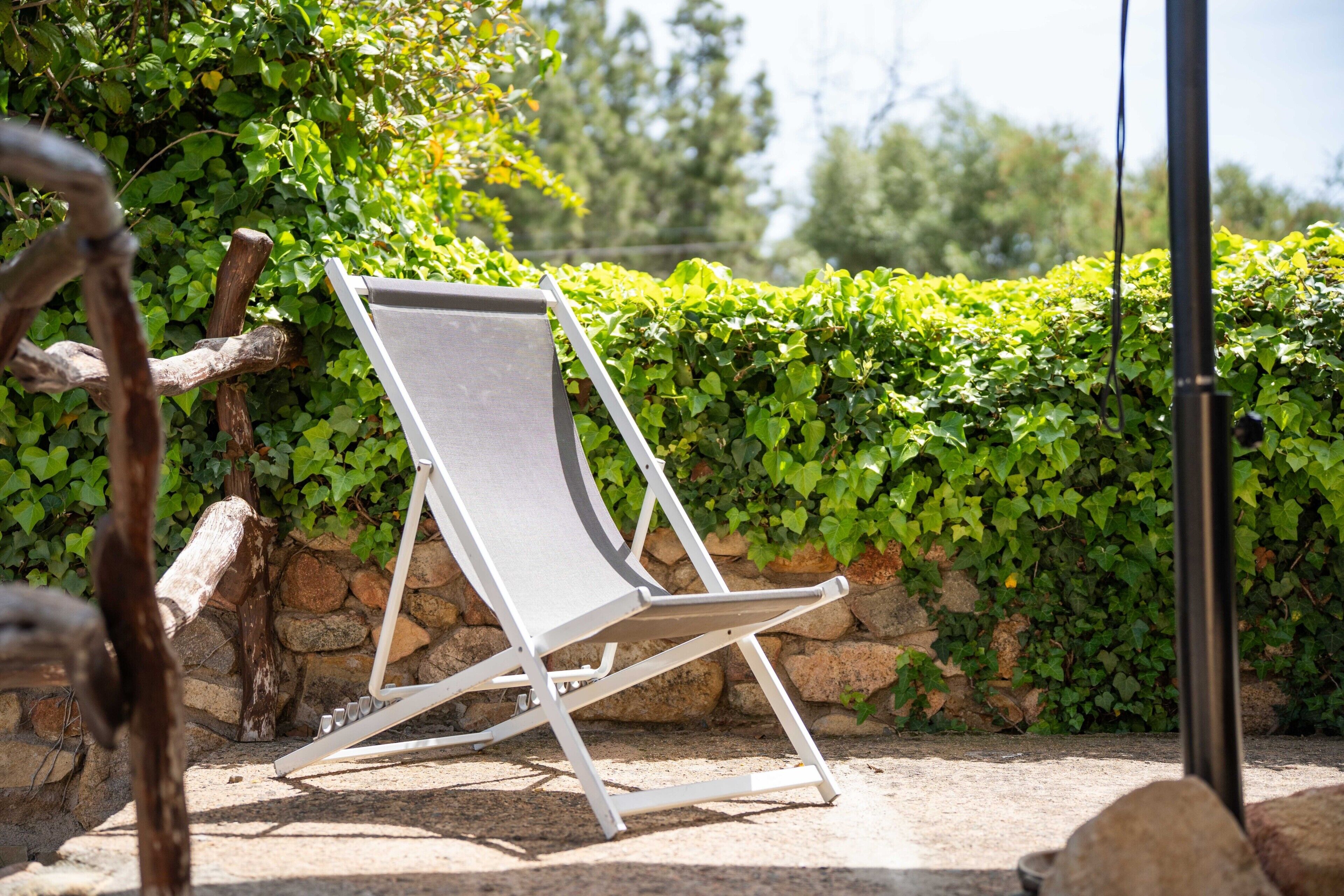 A white lounge chair with a metal frame, positioned outdoors with a stone wall and green foliage in the background.