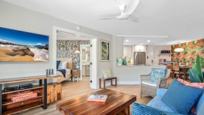 A modern living room with a wooden coffee table, a sofa with blue cushions, a dining area with wicker chairs, and a kitchen with a refrigerator.
